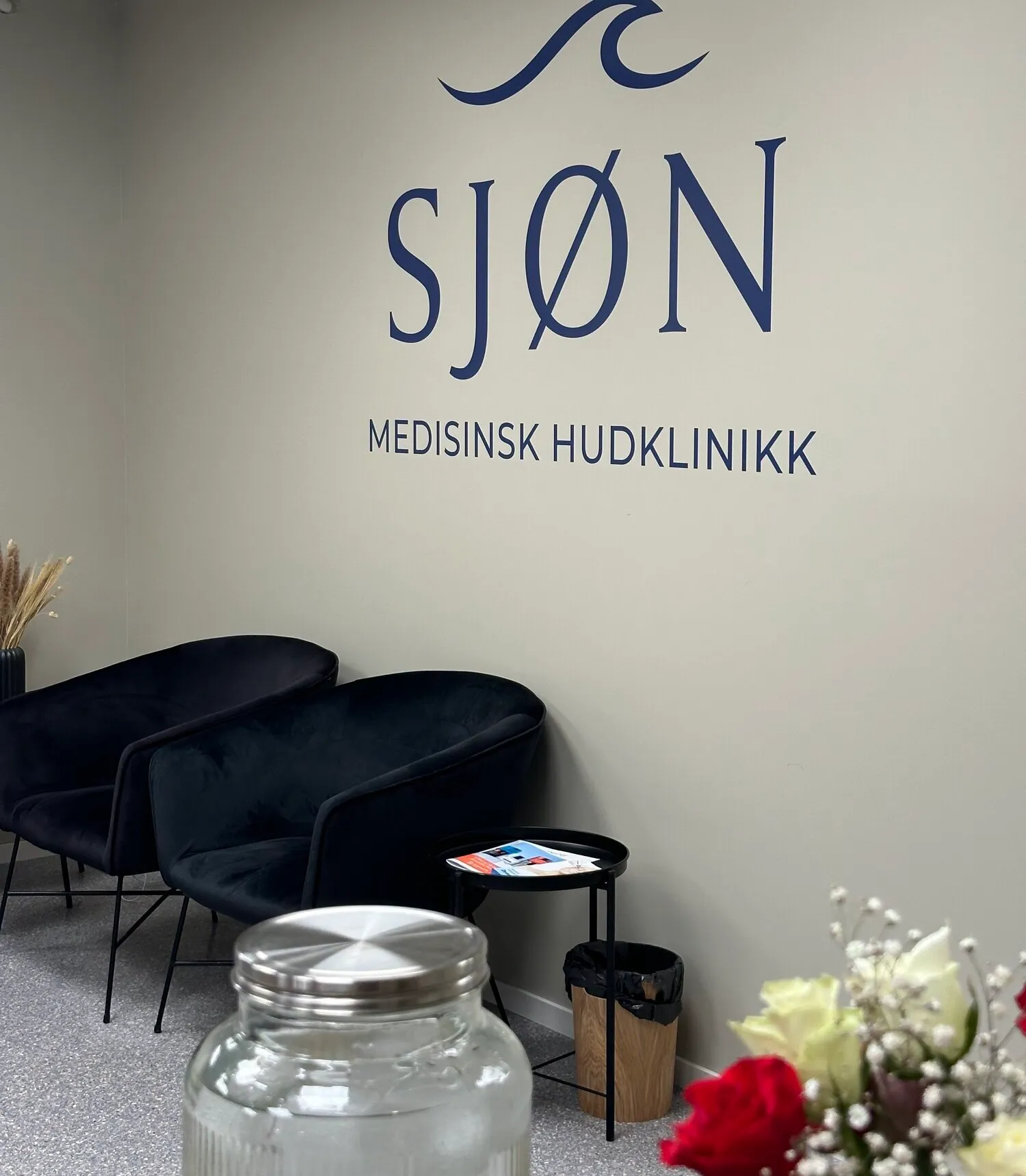 Venteområde hos Sjøn med Sjøn-logo på veggen, lenestoler og friske blomster.